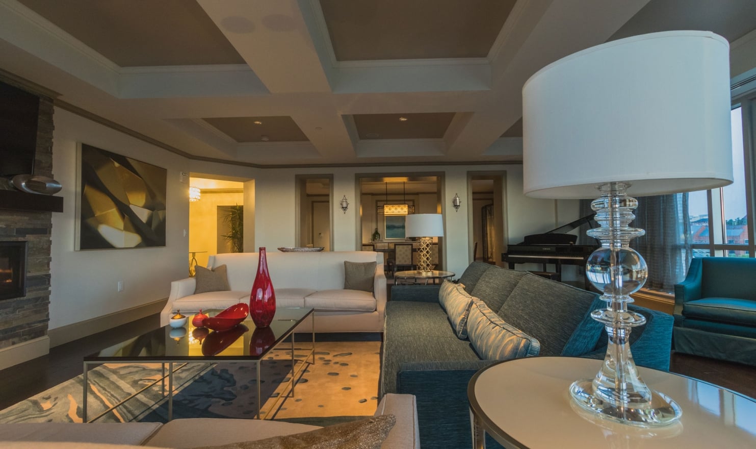 A luxurious living room features beige and teal sofas, a glass coffee table with red vases, and a Yamaha grand piano, in a spacious interior with decorative lighting and large windows.
