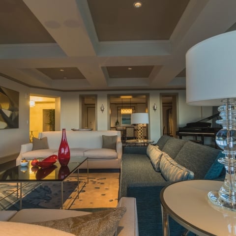 A luxurious living room features beige and teal sofas, a glass coffee table with red vases, and a Yamaha grand piano, in a spacious interior with decorative lighting and large windows.