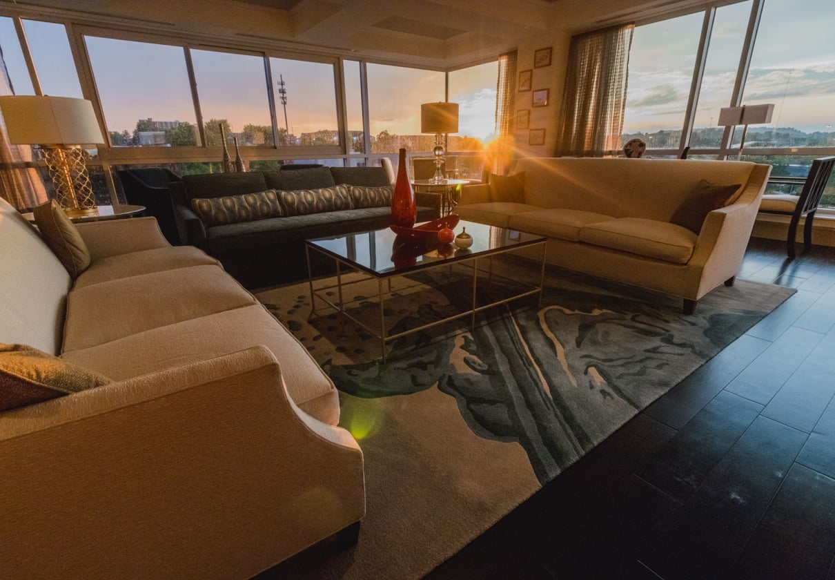 Luxurious living room featuring multiple sofas and a glass coffee table. It's bathed in golden light from a sunset streaming through large windows overlooking a distant cityscape.