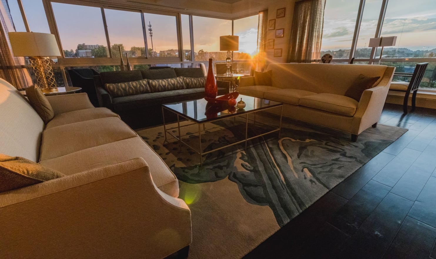 Luxurious living room featuring multiple sofas and a glass coffee table. It's bathed in golden light from a sunset streaming through large windows overlooking a distant cityscape.