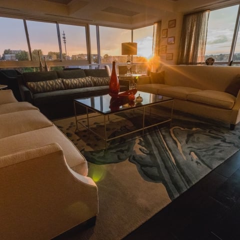 Luxurious living room featuring multiple sofas and a glass coffee table. It's bathed in golden light from a sunset streaming through large windows overlooking a distant cityscape.