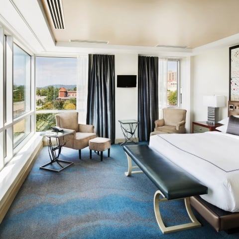 A modern hotel bedroom is furnished with a bed, seating area, and large windows overlooking a city park. A nautical chart featuring "CUMBERLAND CHANNEL LINE" is on the wall.