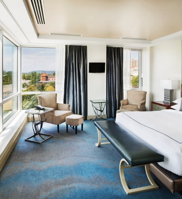 A modern hotel room, with a bed facing expansive city views through floor-to-ceiling windows. It features two armchairs, side tables, and a large map on the wall.
