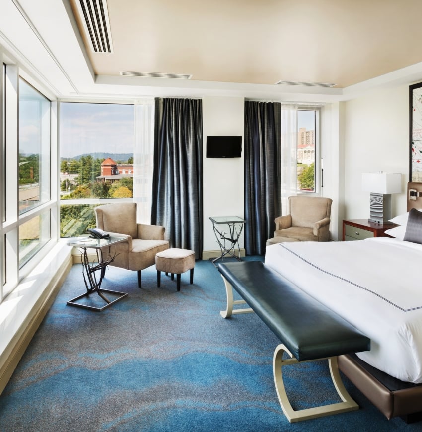 A modern hotel room, with a bed facing expansive city views through floor-to-ceiling windows. It features two armchairs, side tables, and a large map on the wall.