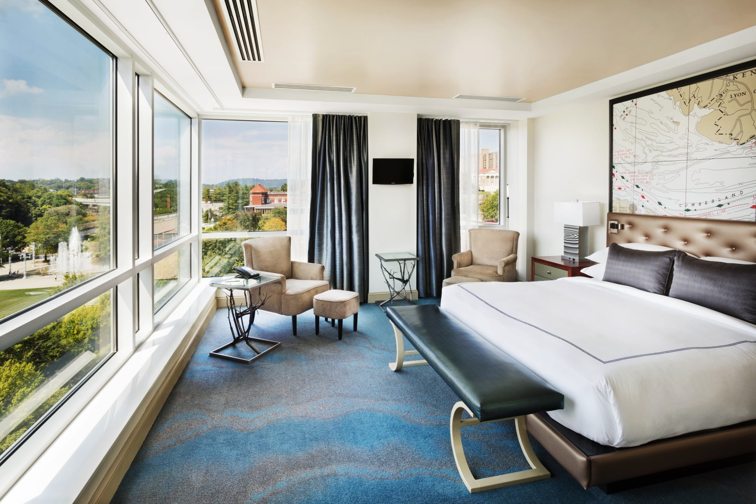 A modern hotel room, with a bed facing expansive city views through floor-to-ceiling windows. It features two armchairs, side tables, and a large map on the wall.