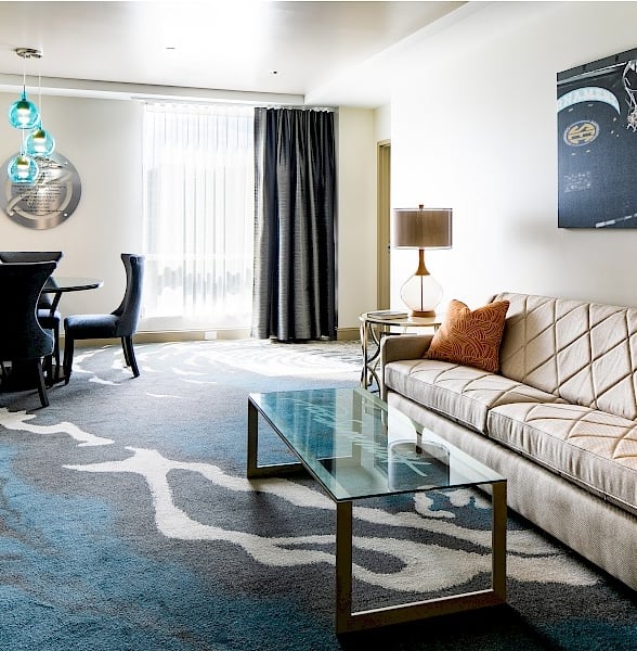 Modern hotel suite features a grey sofa, glass coffee table, and dining set. Arranged in a brightly lit room with a patterned carpet.