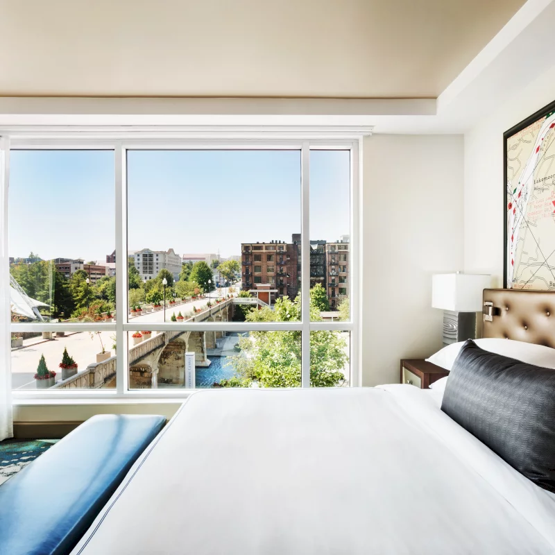 A modern bedroom featuring a white bed and blue bench overlooks a sunny city through a large window. A river, stone bridge, and buildings are visible. Map text: CHATTANOOGA, Lookout Mountain, Missionary Ridge, Chickamauga.