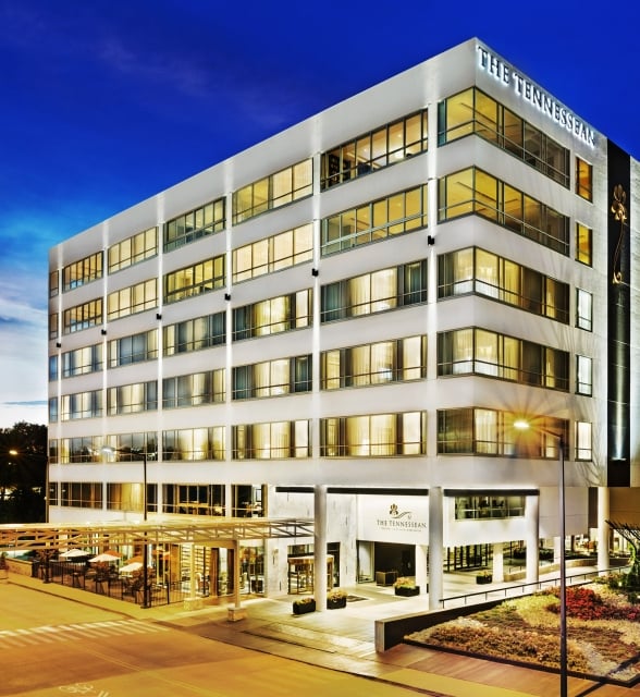 The multi-story white building, "THE TENNESSEAN", glows from internal lights at dusk. It stands by a street, outdoor dining patio, and landscaped gardens, under a blue twilight sky.