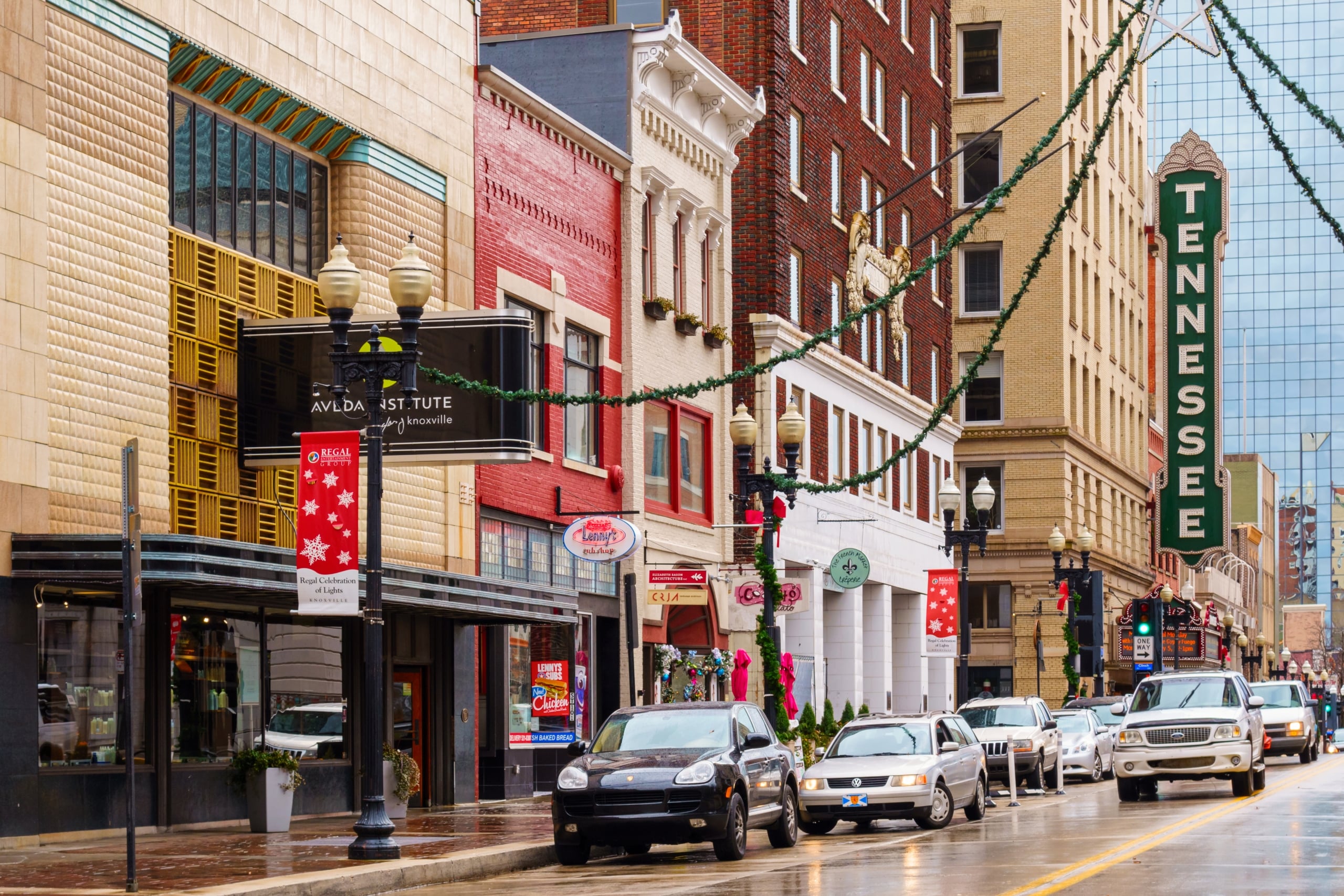 A festive city street features garland-draped lampposts, cars on wet pavement, and buildings of varied architecture. Signs read "TENNESSEE" and "Regal Celebration of Lights Knoxville."