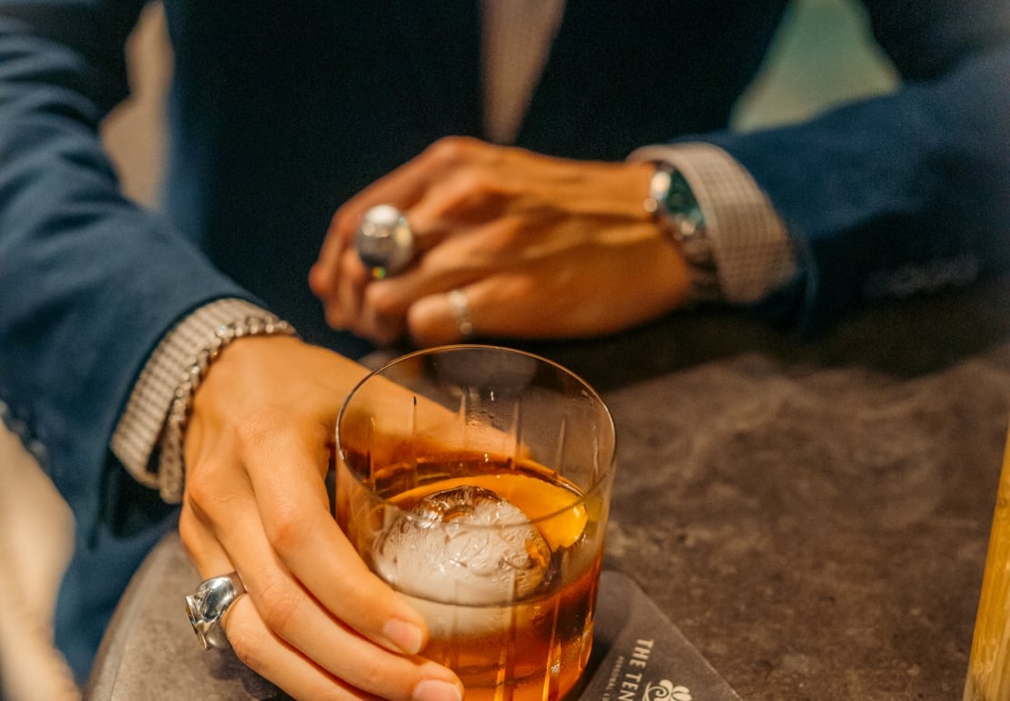 A close-up of a person in a blue blazer holding an amber cocktail with a round ice sphere at a table.