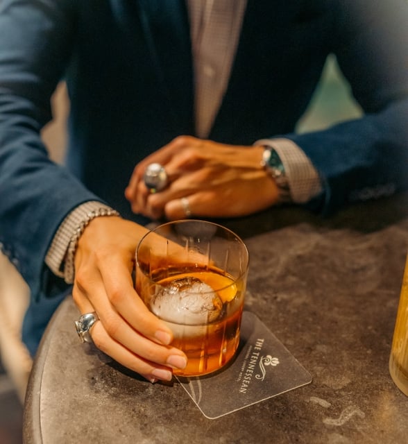A close-up of a person in a blue blazer holding an amber cocktail with a round ice sphere at a table.