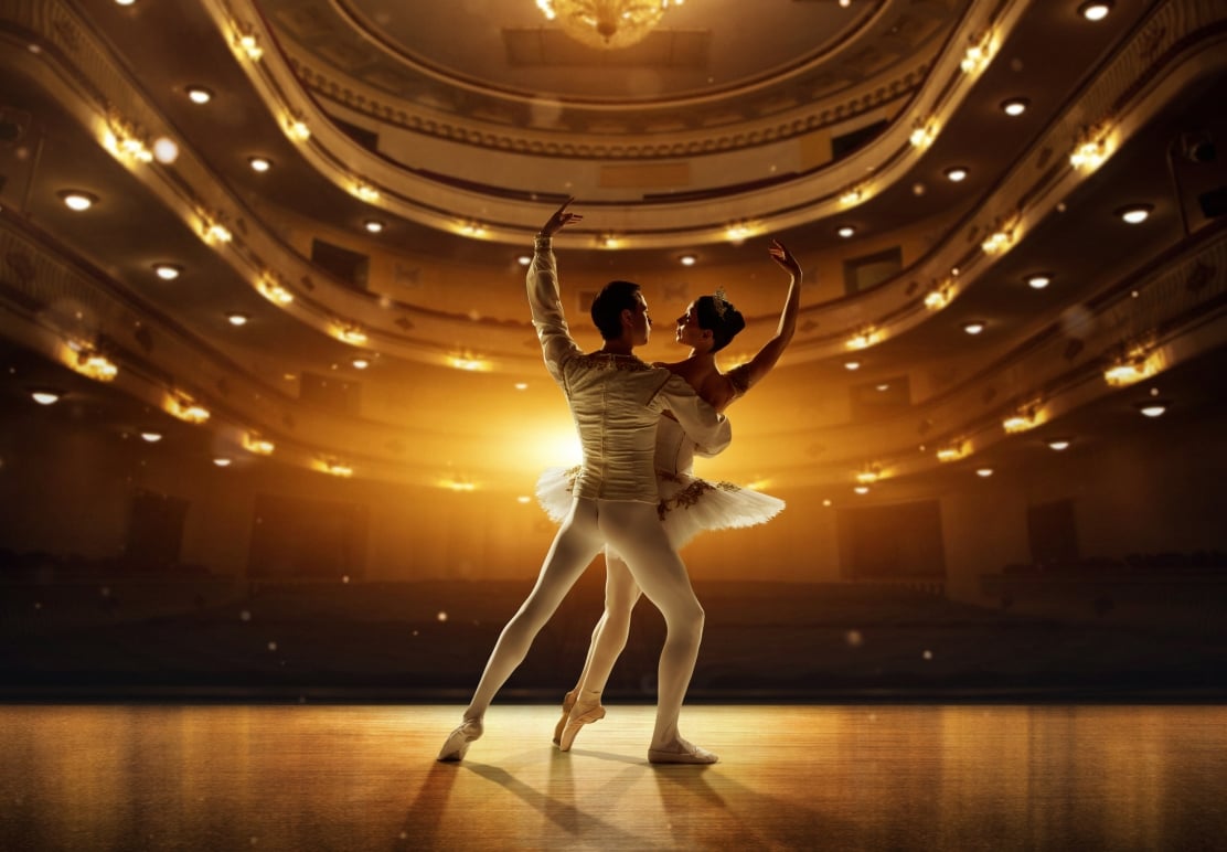 A male and female ballet couple performs a graceful pose on a brightly lit wooden stage, against the backdrop of an ornate, illuminated theater balcony.