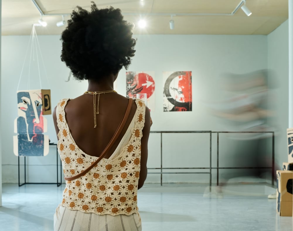 A person with an Afro, seen from behind, stands viewing abstract art in a brightly lit gallery featuring wall paintings and suspended/stacked cardboard sculptures.