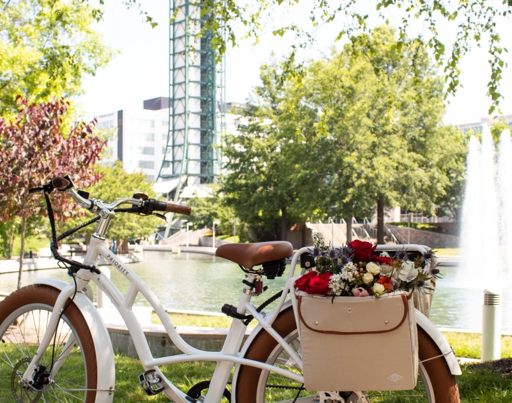 A white "PRIORITY THE TENNESSEAN" bicycle, featuring a "174" canvas bag of flowers, is parked by a pond with a fountain, trees, and a tall tower in sunlight.