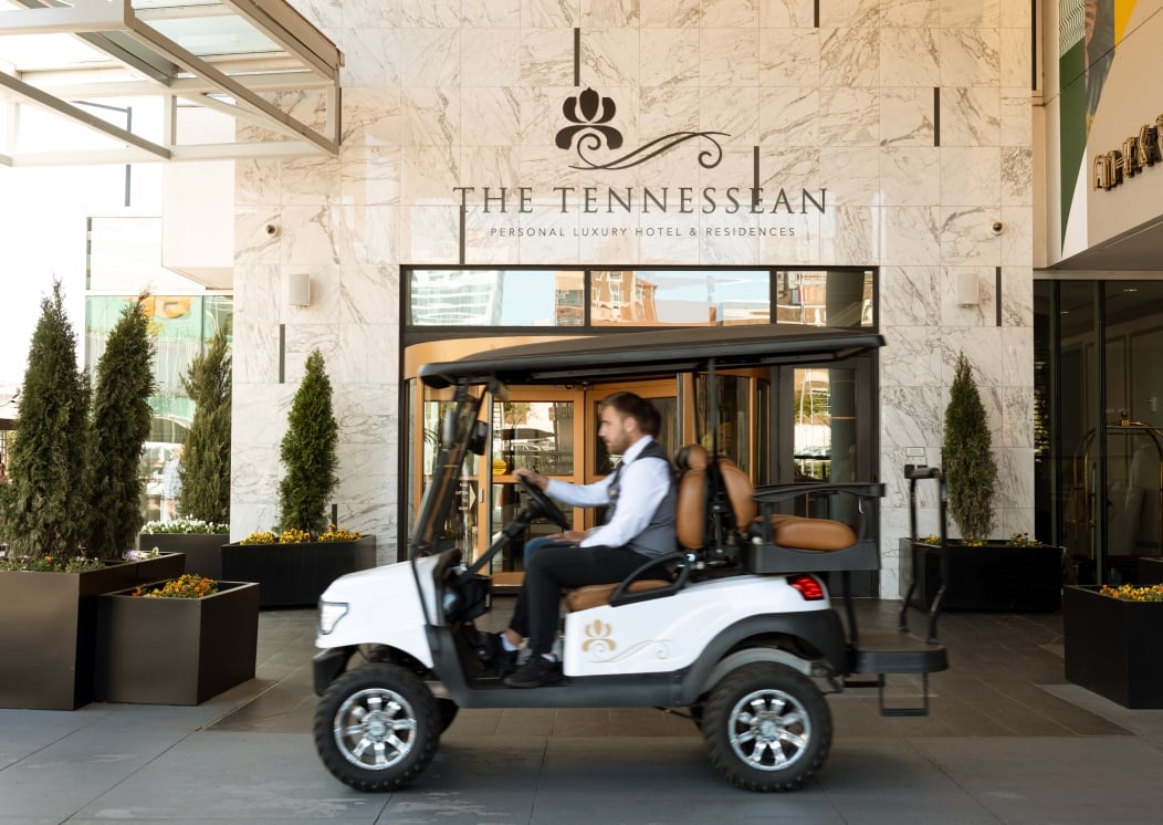 A man drives a white golf cart past a marble-fronted building. Text: THE TENNESSEAN PERSONAL LUXURY HOTEL & RESIDENCES.