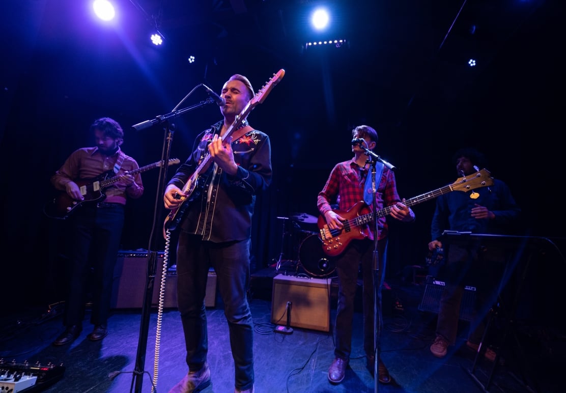 A four-piece band performs on a dimly lit stage under blue lighting. The lead singer/guitarist plays intently, flanked by a guitarist, bassist, and another musician playing keyboard/percussion.
