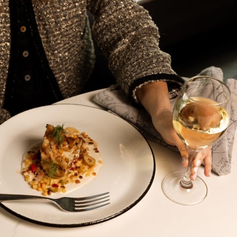 A person's hand in a sparkly jacket holds a white wine glass beside a gourmet plated meal of fish, sauce, and grains on a marble table.