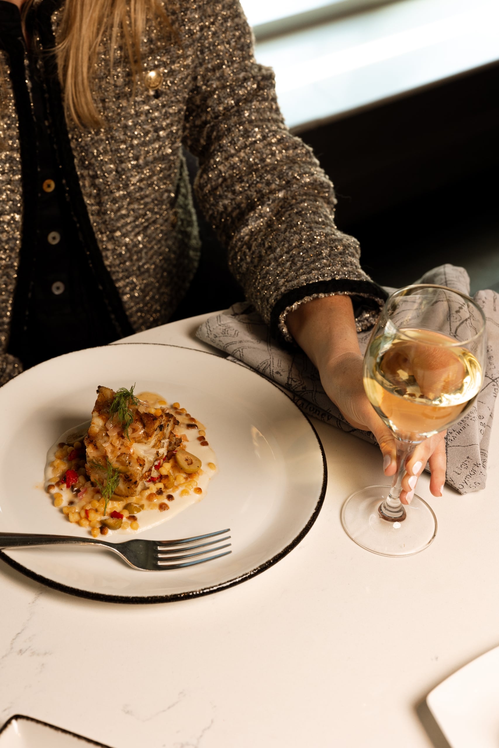 A person's hand in a sparkly jacket holds a white wine glass beside a gourmet plated meal of fish, sauce, and grains on a marble table.
