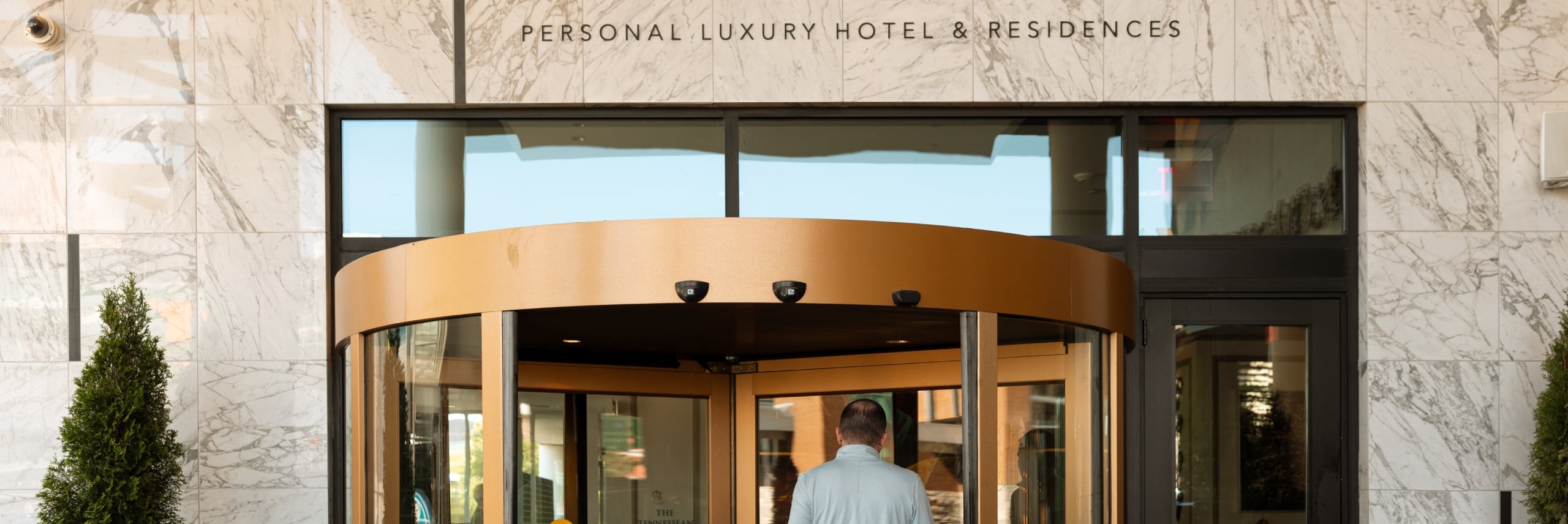 A rear view of a man carrying a leather briefcase, walking through a revolving glass door into a hotel lobby.