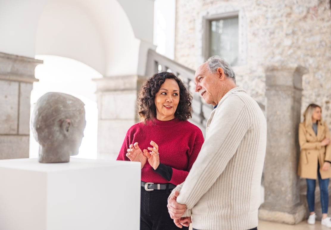 Two people converse, looking at a bust sculpture on a white pedestal, inside a stone-walled art gallery. The woman in red gestures while speaking.
