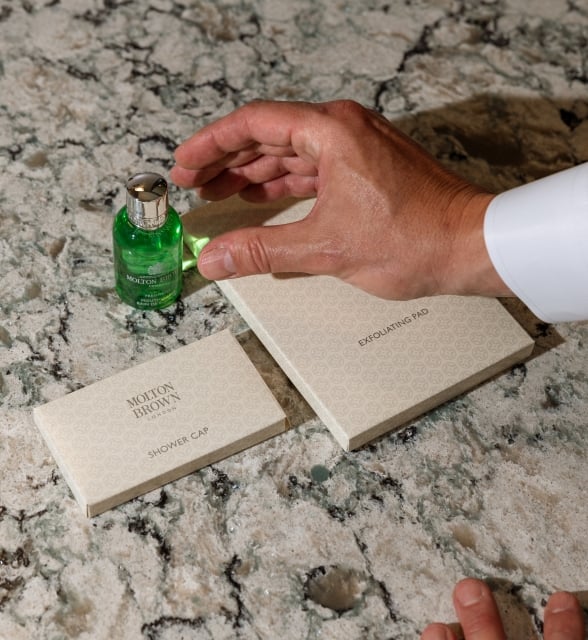 A close-up shot of a hand placing a green bottle of Molton Brown body wash on a marble countertop next to a shower cap.