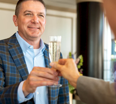 A smiling man in a blue plaid suit clinks a champagne flute with another person's hand, celebrating in an indoor setting.