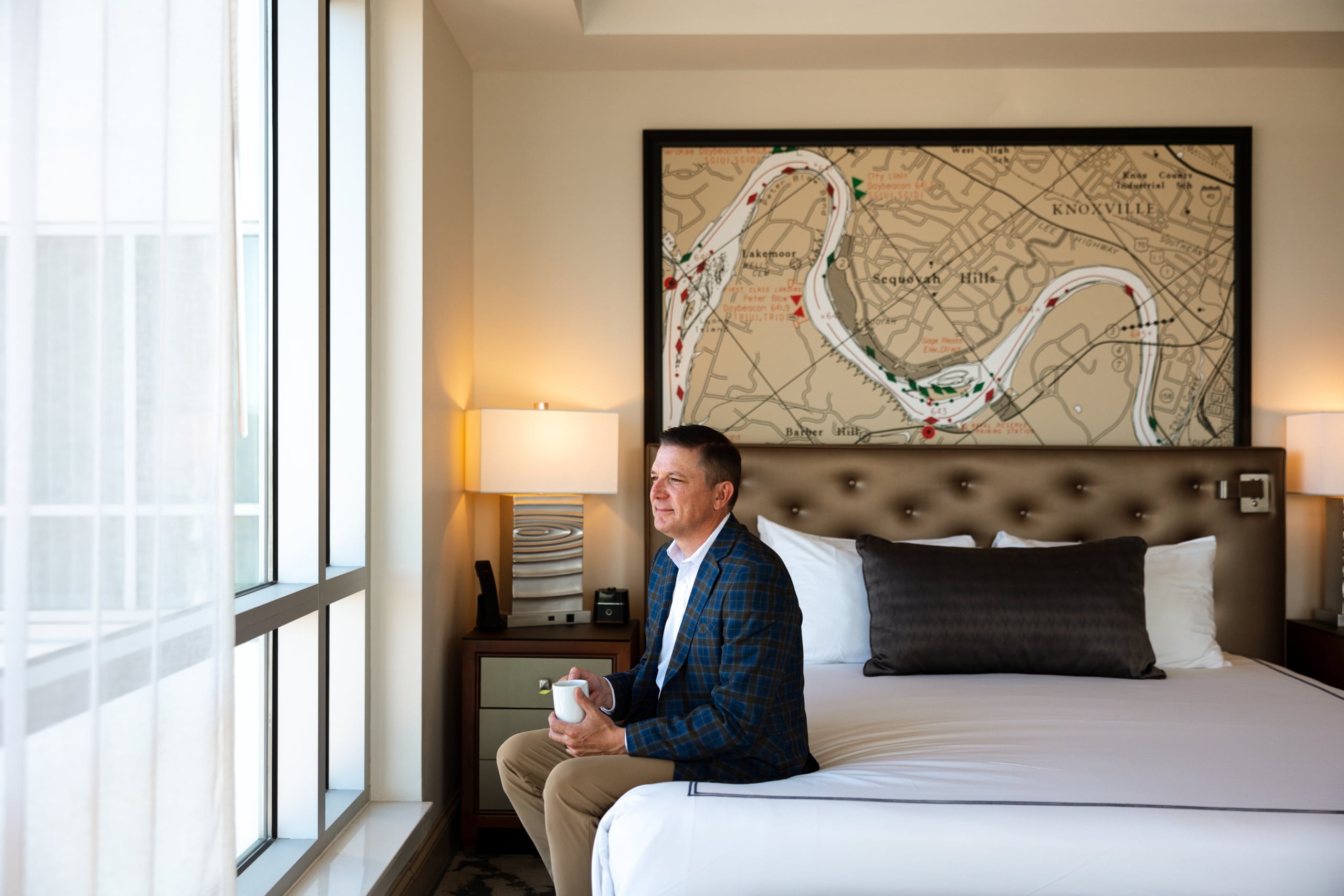 Man in a plaid jacket sits on a bed's edge, holding a mug and looking out a window. A map of "KNOXVILLE" hangs above the bed.