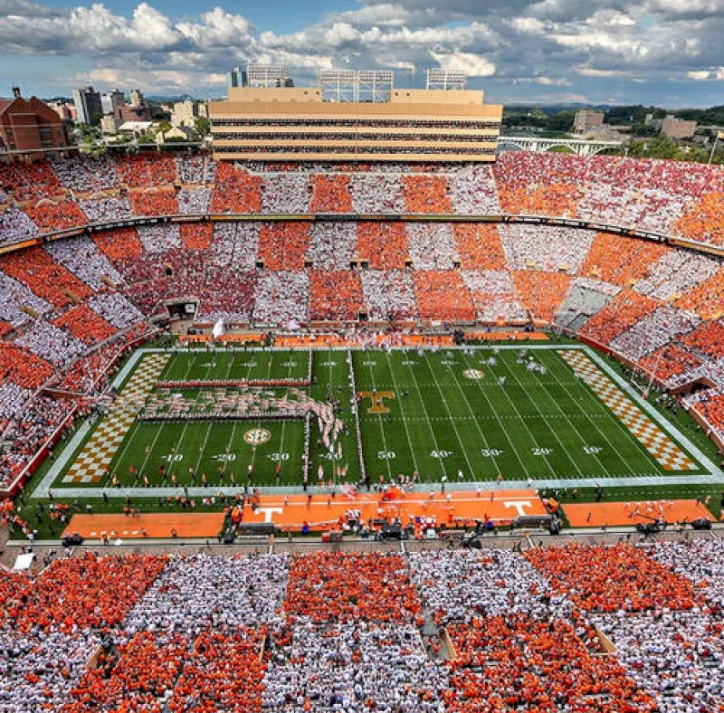 A packed football stadium features fans in orange and white forming checkerboard patterns. Players are on the green field marked with a large "T" and "SEC" logos, surrounded by city buildings under a cloudy sky.