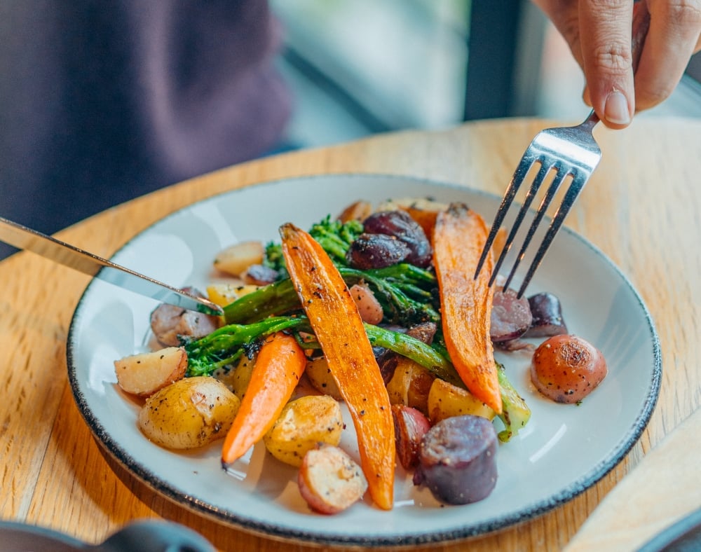 A close-up shot of a person using a fork to pick up roasted vegetables, including carrots and potatoes, from a plate.