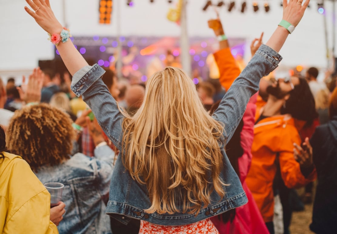Blonde-haired person, seen from behind, raises arms in celebration amidst a cheering crowd at a lively outdoor concert, with blurry stage lights.