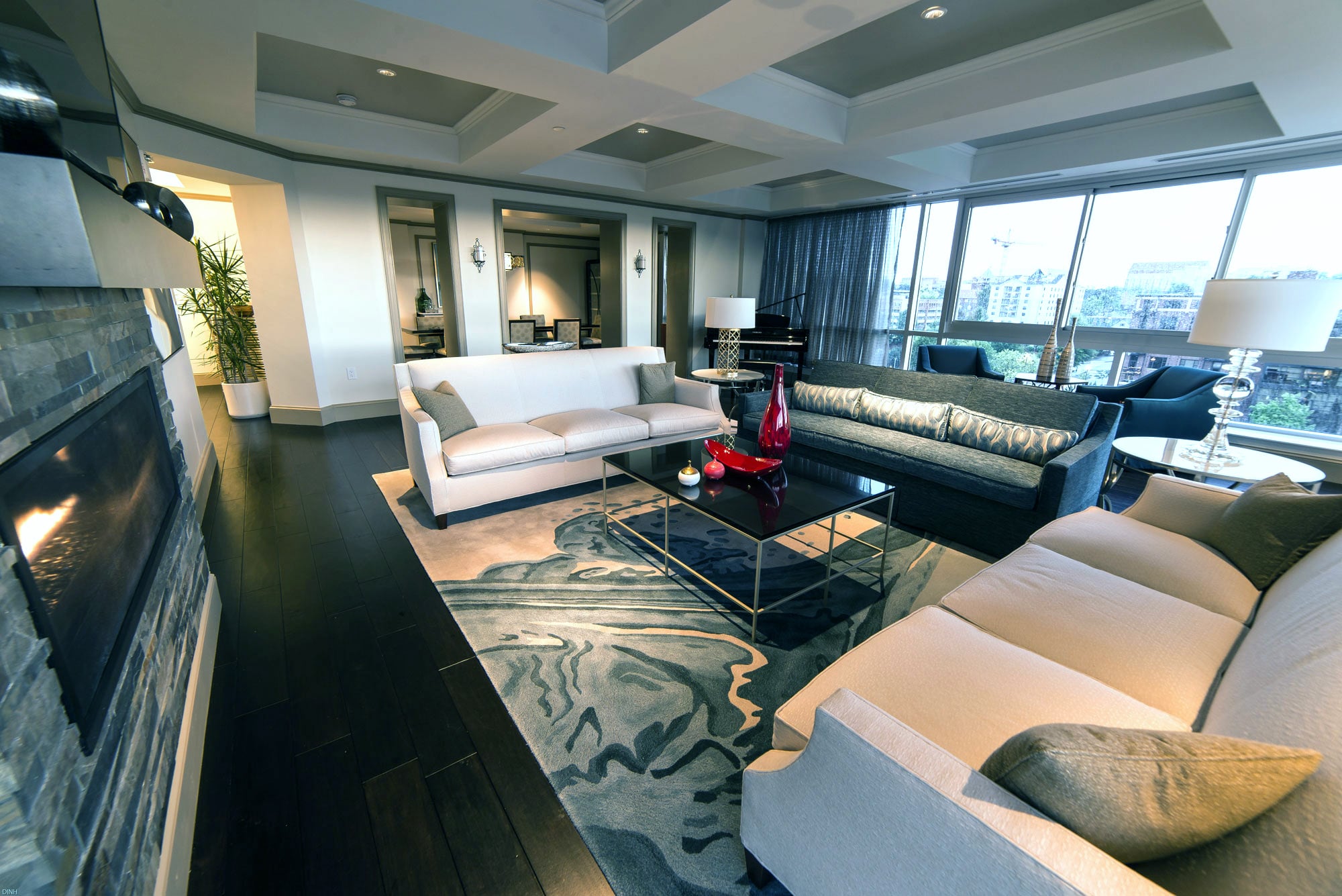 Modern living room: white and teal sofas, coffee table with red decor, fireplace, and a Yamaha grand piano near windows with a city view. Dark floors, abstract rug.