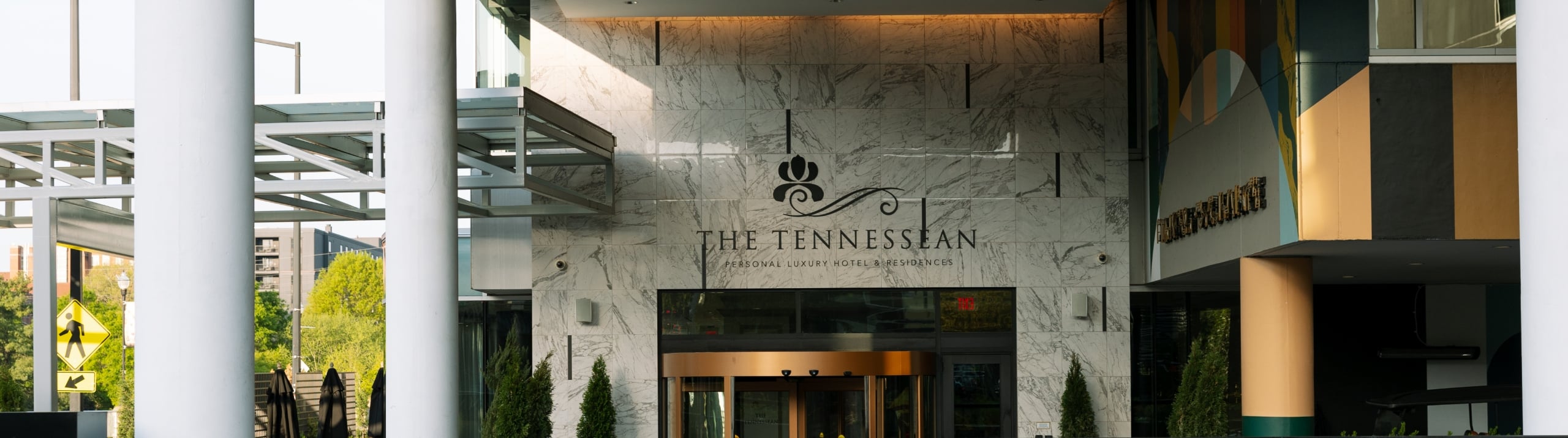 The Tennessean hotel entrance features a large revolving door set into a marble facade, framed by white pillars, with green plants in the foreground. Text: THE TENNESSEAN PERSONAL LUXURY HOTEL & RESIDENCES.