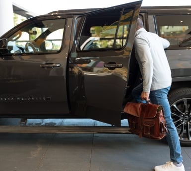 A man carrying a brown leather briefcase steps into a dark grey SUV parked outside a building.