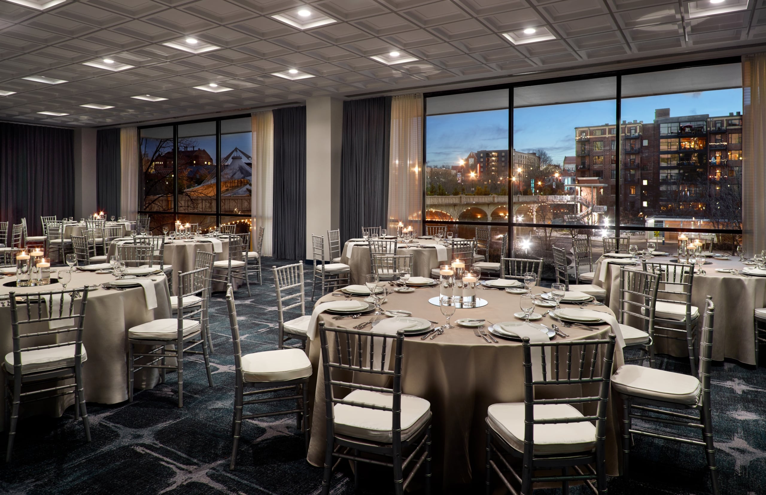 Round tables are formally set with place settings and candle centerpieces, surrounded by silver chairs, in a large event room with city views and a bridge at dusk.