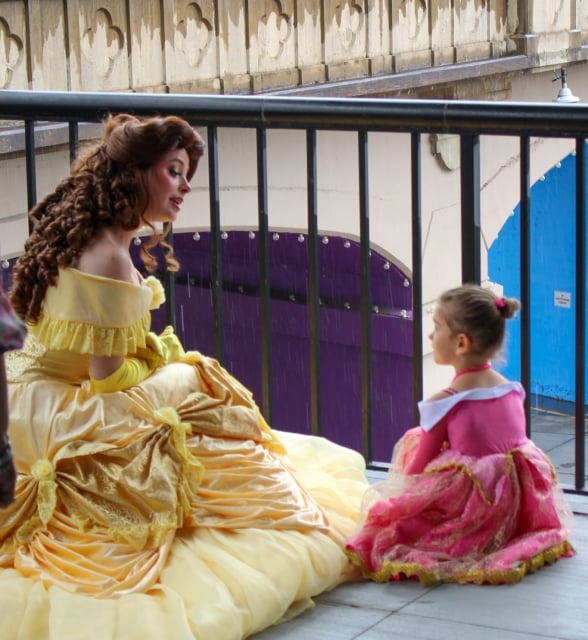 A woman dressed as Belle from Beauty and the Beast talks to a young girl in a pink princess dress outdoors.