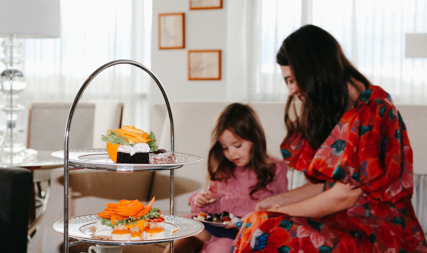 A two-tiered stand displays colorful desserts, including orange-rose garnishes. In the background, a woman and child enjoy small treats at a bright indoor table.