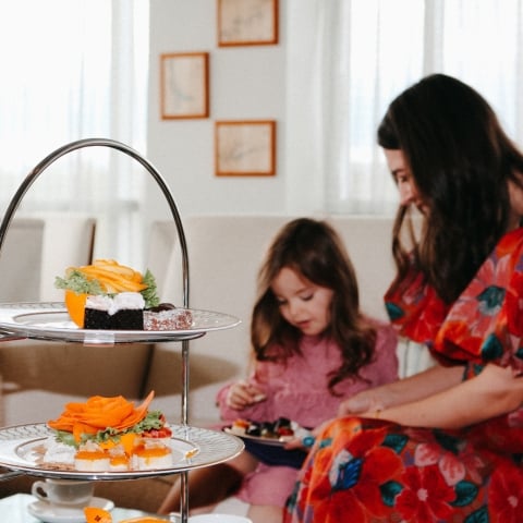 A two-tiered stand displays colorful desserts, including orange-rose garnishes. In the background, a woman and child enjoy small treats at a bright indoor table.