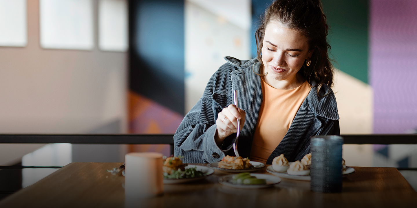 A woman smiles while eating from a plate with a fork at a wooden table, in a colorful restaurant with geometric wall designs.