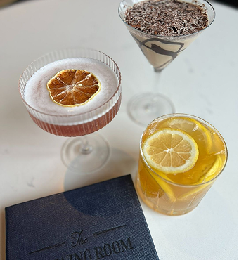 Three distinct cocktails and a blue menu sit on a white table. One is a martini with chocolate shavings, another a coupe glass with a dried orange, and the third a rocks glass with lemon slices. Text: The Drawing ROOM.