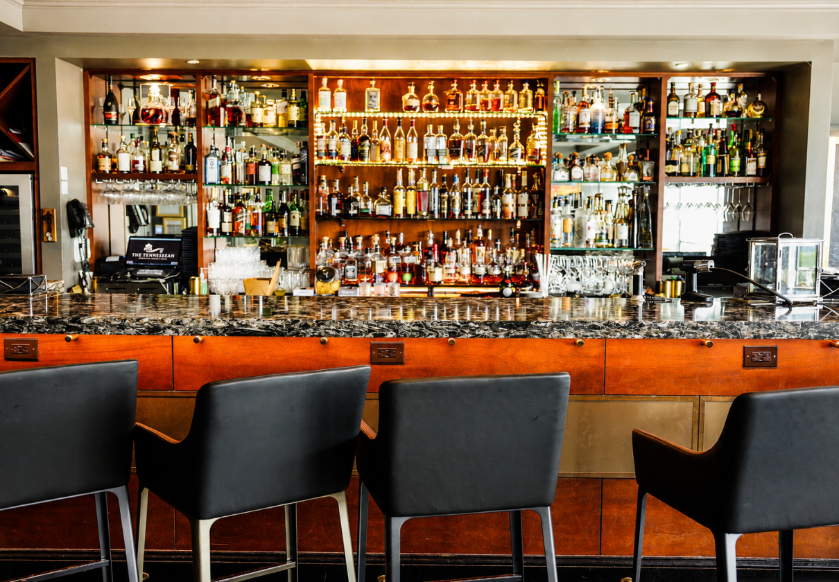 An upscale bar with black chairs, a granite countertop, and rows of liquor bottles on illuminated shelves.