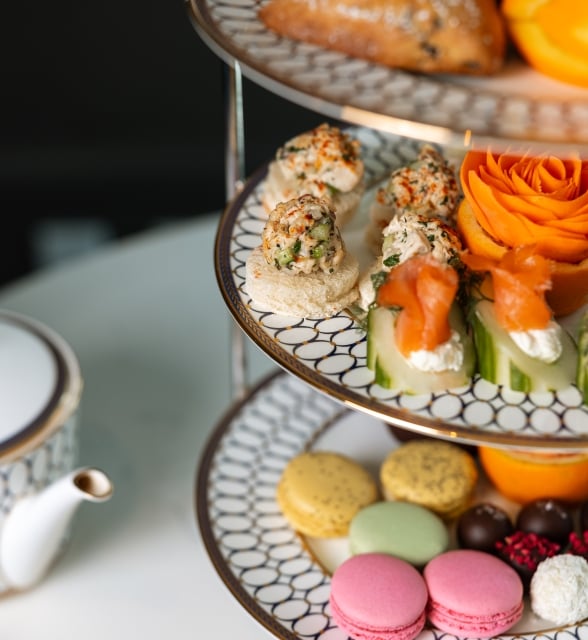 A three-tiered high tea tray filled with pastries, macarons, and sandwiches next to a patterned white teapot.