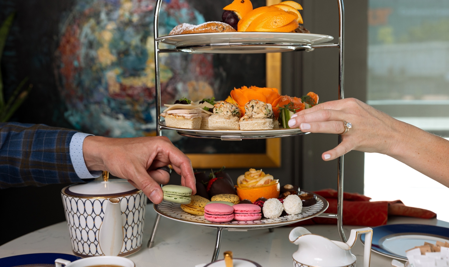 A man and woman reach for varied sandwiches, pastries, and macarons on a three-tiered stand at a tea party, surrounded by elegant tableware and abstract art.