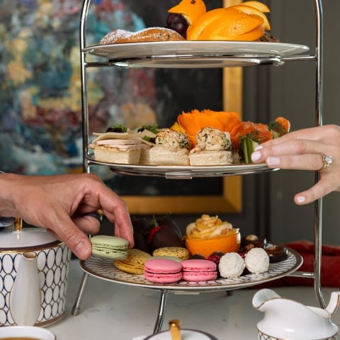 A man and woman reach for varied sandwiches, pastries, and macarons on a three-tiered stand at a tea party, surrounded by elegant tableware and abstract art.