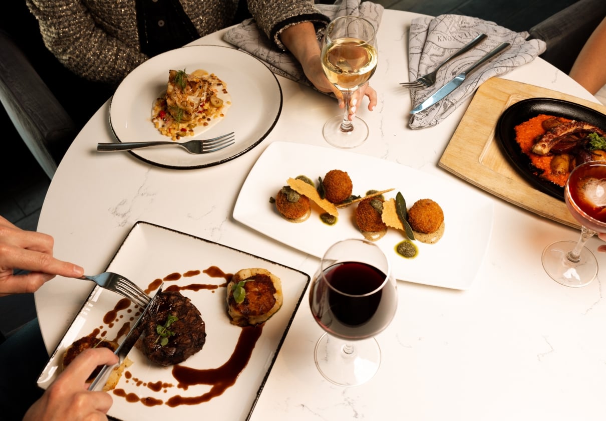 Gourmet dishes and drinks are arranged on a white marble table, being enjoyed by multiple diners. Hands hold cutlery and glasses, with "KNOXVILLE" visible on a napkin.