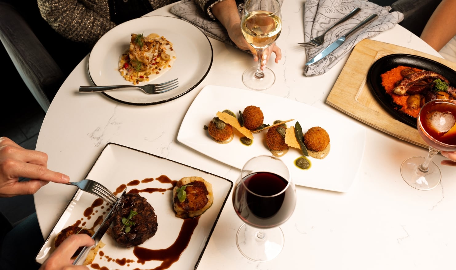 Gourmet dishes and drinks are arranged on a white marble table, being enjoyed by multiple diners. Hands hold cutlery and glasses, with "KNOXVILLE" visible on a napkin.