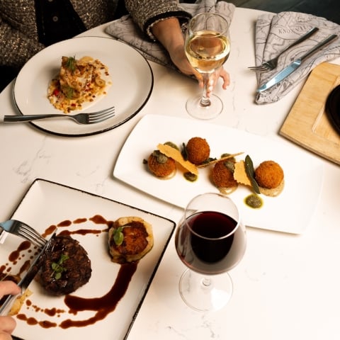 Gourmet dishes and drinks are arranged on a white marble table, being enjoyed by multiple diners. Hands hold cutlery and glasses, with "KNOXVILLE" visible on a napkin.