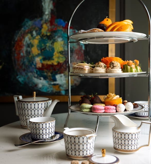 A three-tiered high tea tray filled with pastries, sandwiches, and fruit next to a patterned white and gold tea set.