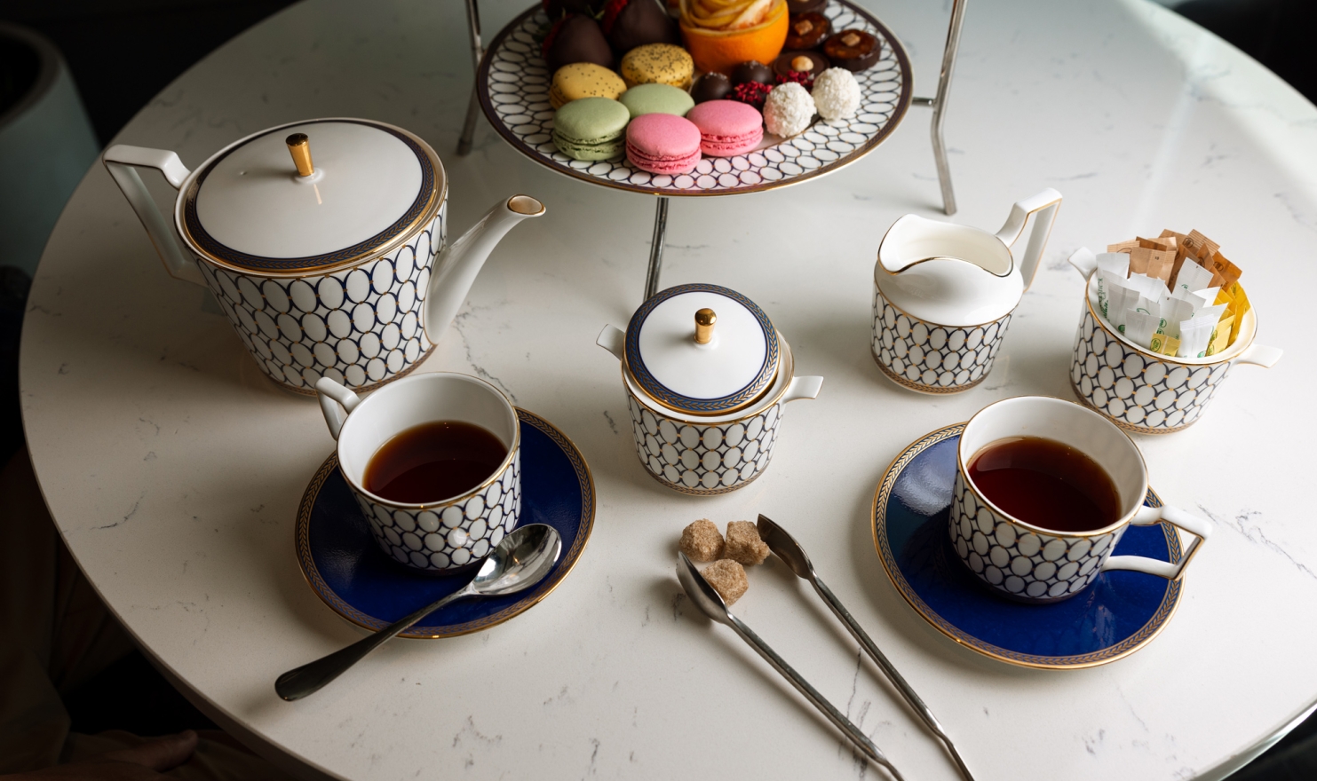 An elegant white and gold patterned afternoon tea set with tea-filled cups, sugar, and creamer is displayed on a white marble table. A tiered stand holds colorful macarons and pastries.