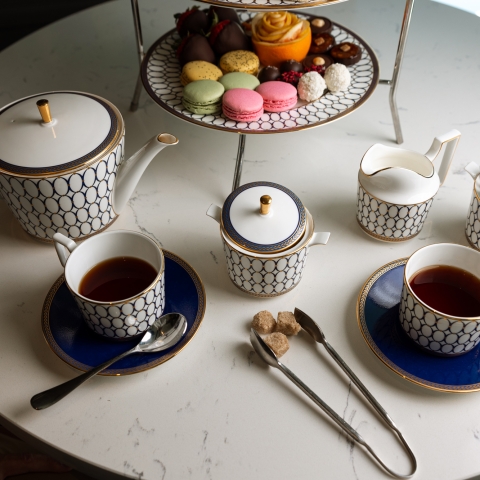 An elegant white and gold patterned afternoon tea set with tea-filled cups, sugar, and creamer is displayed on a white marble table. A tiered stand holds colorful macarons and pastries.