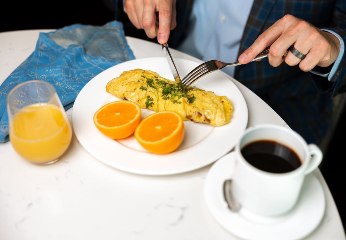 A person's hands, wearing a ring, cut into a herb-garnished omelet alongside orange halves, orange juice, and black coffee on a white table.